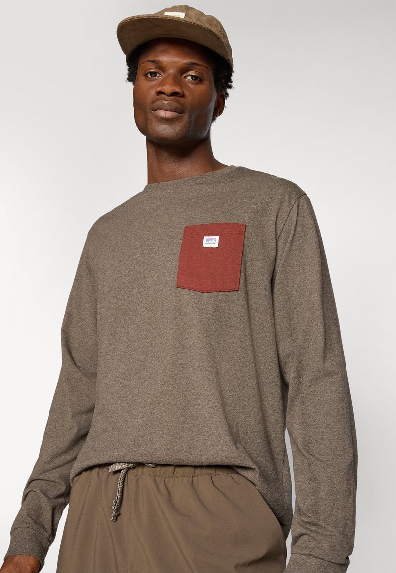 Langearmige bruine shirt met een bordeauxrood borstzakje, voorzien van een klein merklabel. Gecombineerd met beige shorts met koord.