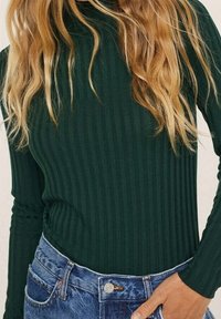 Frau in einem gerippten dunkelgrünen Langarmoberteil, das in hochgeschnittene blaue Jeans gesteckt ist, mit einer Hand, die in der Nähe der Tasche ruht.