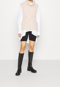 Beiges, gestricktes, ärmelloses Rollkragensweatshirt über einem weißen Hemd, kombiniert mit schwarzen Fahrradhosen und knielangen schwarzen Lederstiefeln.