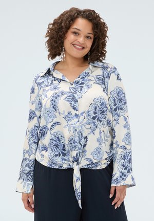 Donna sorridente con capelli ricci che indossa una camicia a fiori blu e bianca abbottonata e annodata in vita e pantaloni scuri, in piedi davanti a uno sfondo semplice.