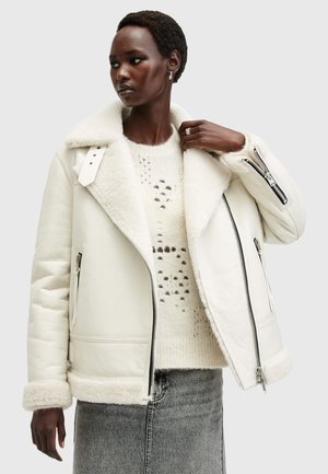 Weiße Lederjacke mit Schurwollkragen und -manschetten, ausgestattet mit zwei Reißverschlüssen und seitlichen Taschen, über einem leichten Pullover getragen.
