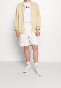 Ljusbeige zip-up-jacka, vit t-shirt med blomsterbroderi, vita shorts och ljusfärgade sneakers. Mjuka texturer, avslappnad passform.