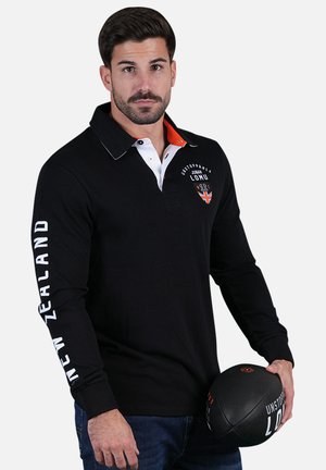 Polo noir à manches longues avec des accents de col orange et du texte blanc "NOUVELLE-ZÉLANDE" sur la manche gauche ; modèle tenant un ballon de football noir.