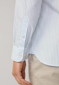 Chemise à manches longues rayée bleu clair et blanche avec un poignet boutonné. Le tissu apparaît lisse avec une coupe décontractée et un ourlet arrondi.