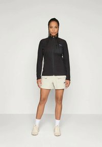 Casaco de fleece preto com fecho, de mangas longas, combinado com calções bege claros. Os detalhes notáveis incluem um logo no peito e bolsos laterais.