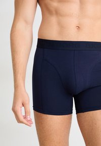 Mees torso ja ülemised jalad, seljas tumesinine boxer-šortid logoga vööga, seismas neutraalse helebackgroundi ees.