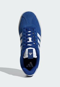 Blå mockasneakers med vita ränder, platta snören, vadderad krage och svart innersula. Har en logotyp på tungan och bakre fliken.