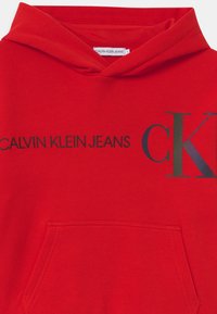 Sudadera de algodón roja con bolsillo frontal, una gran impresión de "CALVIN KLEIN JEANS", y un logotipo "CK" en contraste en el pecho.