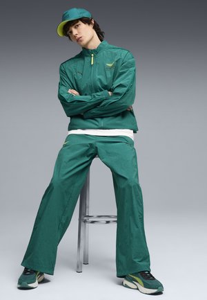 Junge Person sitzt auf einem Hocker, trägt einen grünen Trainingsanzug, eine passende Mütze und Turnschuhe vor einem grauen Hintergrund.