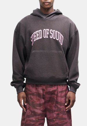 Uomo che indossa una felpa con cappuccio grigio scuro con testo "SPEED OF SOUND" e pantaloni larghi rossi con fantasia su sfondo neutro.