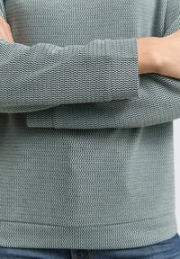 Light green oversized sweatshirt with a textured zigzag pattern, featuring long sleeves and a relaxed fit, paired with denim.