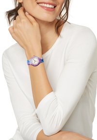 Farverig ur med et rundt rem i lilla og blå m�ønster, en hvid urskive og timemarkeringer i guldton, båret på en lys hudfarve håndled.