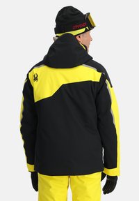 Schwarze und gelbe Skijacke mit Kapuze, die ein markantes, schräges gelbes Panel und reflektierende Akzente aufweist. Enthält ein Spinnenlogo auf der Rückseite.