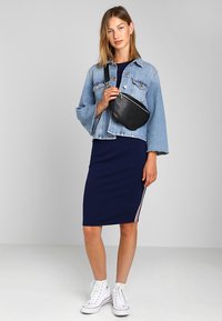 Giacca di denim con maniche sfilacciate, indossata su un abito bodycon blu navy con strisce laterali rosse e bianche, abbinato a sneakers alte bianche.