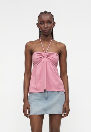 Femeie cu părul împletit, purtând un top halter roz și o fustă mini din denim bleu deschis, stând în fața unui fundal alb simplu.