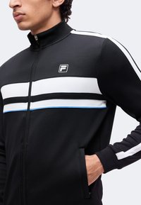 Homme portant une veste noire zippée avec des rayures blanches horizontales, petit logo sur la poitrine et main dans la poche sur fond uni.