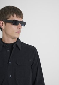Camisa de pana negra con dos bolsillos frontales, cierre de botones y una sutil raya de acento horizontal; combinada con gafas de sol rectangulares oscuras.