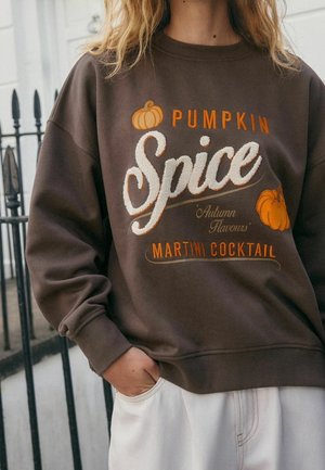 Personne portant un sweat-shirt marron avec le texte « Pumpkin Spice Autumn Flavours Martini Cocktail » et des illustrations de citrouilles. Main dans la poche d'un pantalon blanc.