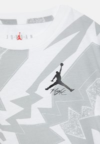 Biała koszulka z abstrakcyjnymi szarymi wzorami błyskawic, z czarnym logo Jumpman i napisem "Flight" pod spodem.