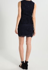 Abito in pizzo blu navy composto da un top senza maniche e una minigonna. Motivi floreali e dettagli con orlo a scallop. Sandali neri con tacco.