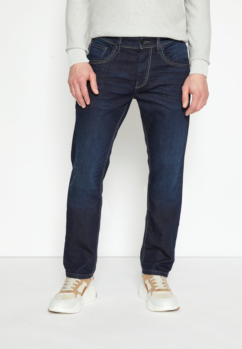 Donkerblauwe denim jeans met contrasterende witte stiksels, mid-rise ontwerp, vijf zakken en een relaxte pasvorm, gecombineerd met beige sneakers.