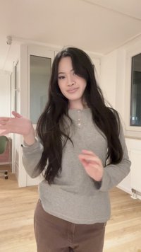 Giovane donna con lunghi capelli scuri che indossa una maglietta grigia e pantaloni marroni, che gesticola con le mani in una stanza moderna con pavimenti in legno.