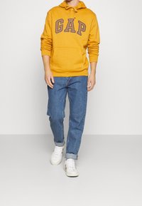 Sudadera de color mostaza con el logo de "GAP" en marrón oscuro, bolsillo tipo canguro y capucha con cordón, combinada con jeans azules holgados y zapatillas blancas.