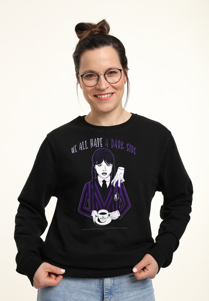Donna sorridente con occhiali che indossa una felpa nera con un personaggio a strisce viola che tiene una tazza con la scritta "Abbiamo tutti un lato oscuro."