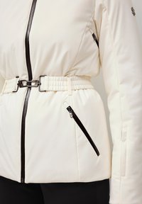 Crème jas met een belted taille, zwarte ritscontrast, gewatteerde textuur en ritszakken, met een gladde afwerking en een getailleerd ontwerp.