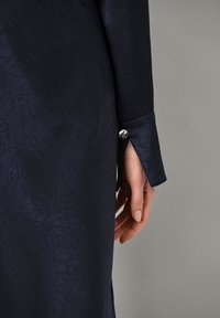 Mörkblå texturerad tygärmsmanchett med silverknappdetalj, som visar ett nötmönster och skräddarsydd ärmslutning.