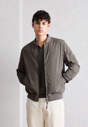 Mladý muž v sivej bomber bunde cez tmavé tričko a biele nohavice, stojaci s rukami vo vreckách pri jednoliatnej stene.