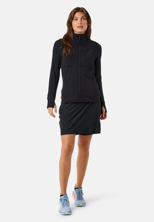 Sort lynlåsejakke med høj krave, letvægtsstof, to forlommer, parret med en sort nederdel og blå atletiske sko.