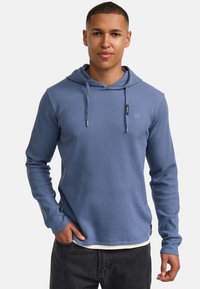 Langärmliges Kapuzenshirt aus strukturiertem blauen Stoff mit einem subtilen Rippenmuster, ausgestattet mit einer Kordelzugkapuze und einem Etikett-Highlight auf der Brust.