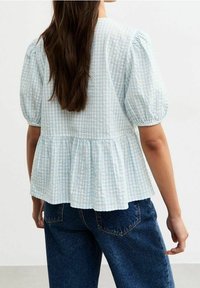 Hellblau kariertes Peplum-Oberteil mit kurzen Puffärmeln und geraffter Taille, aus einem weichen strukturierten Stoff gefertigt. Kombiniert mit dunkelblauen Jeans.