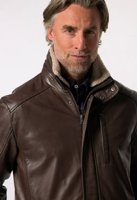 Chaqueta de cuero marrón con cuello de borreguillo, cierre de cremallera y bolsillo lateral. Textura suave y ajuste entallado visible.