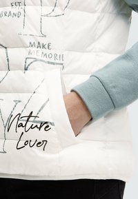 Weiß gesteppte Pufferweste mit grauem Grafiktext "MAKE MEMORIES" und "Nature Lover". Verfügt über eine Fronttasche und einen leicht blauen Ärmelbund.