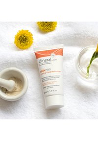 Hidratante Facial Calmante SkinPro en un tubo blanco con acentos naranjas, colocado sobre una toalla blanca con flores amarillas y un mortero y mano de mortero.