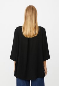 Schwarzes, übergroßes Oberteil aus strukturiertem Stoff, mit weiten Ärmeln und abgerundetem Rückensaum, kombiniert mit blauen Denim-Hosen.