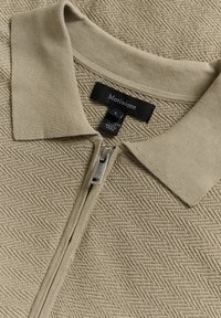 Beige polo s zipem, s texturovaným šikmým vzorem. Obsahuje klasický límeček a stříbrný zip vpředu. Značková etiketa viditelná uvnitř.