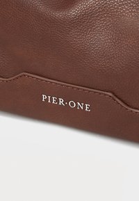 Brunet teksturert lær clutch-veske med en bølgete søm og sølv "PIER ONE"-logo. Glatt overflate og subtil glans.