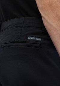 Κοντινό πλάνο της τσέπης μαύρου παντελονιού Calvin Klein στο ισχίο ενός ατόμου, εμφανίζοντας την ετικέτα της μάρκας και ένα μέρος του χεριού τους.