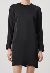 Frau in einem lockeren, langärmeligen schwarzen Satin-Minikleid mit breiten Manschetten und Rundhalsausschnitt vor einem schlichten Hintergrund.