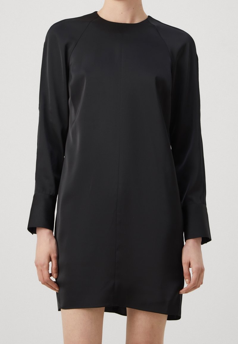 Frau in einem lockeren, langärmeligen schwarzen Satin-Minikleid mit breiten Manschetten und Rundhalsausschnitt vor einem schlichten Hintergrund.