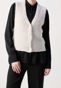 Frau in einem cremefarbenen strukturierten gestrickten Knopfveste über einem schwarzen Langarmhemd mit Kragen und schwarzen Hosen, steht vor einem weißen Hintergrund.