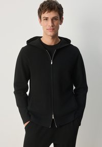 Hanorac negru cu fermoar, dintr-un material moale și texturat, cu glugă căptușită și detaliu cu fermoar argintiu. Purtați peste un tricou negru, design minimalist.