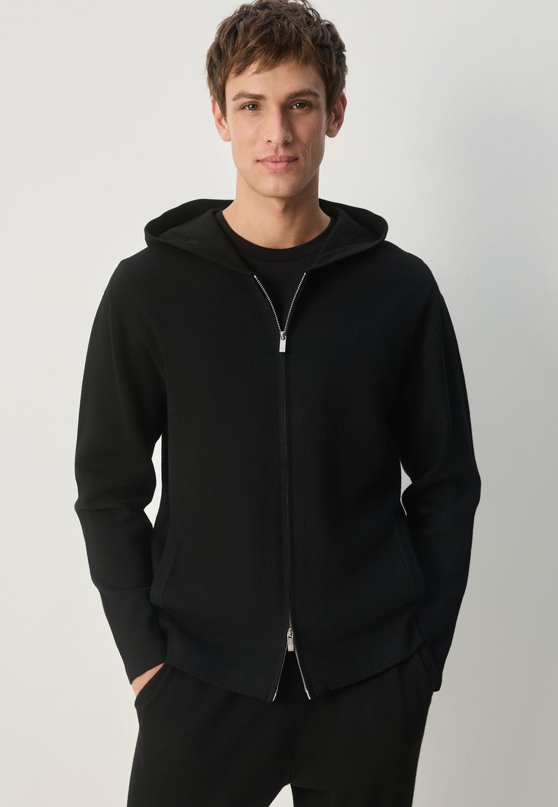 Hanorac negru cu fermoar, dintr-un material moale și texturat, cu glugă căptușită și detaliu cu fermoar argintiu. Purtați peste un tricou negru, design minimalist.