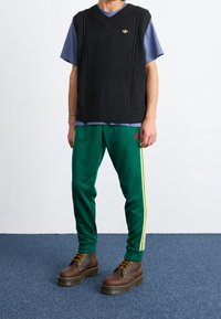 Svart stickad ärmlös tröja, blå T-shirt under, gröna träningsbyxor med sidogula ränder, bruna snörstövlar. Guldig logodetalj.