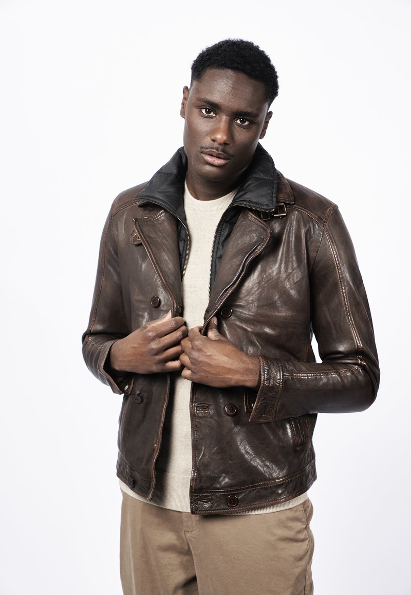 Chaqueta de cuero marrón con cuello negro; diseño de doble botonadura, superficie texturizada, botones oscuros, llevada sobre un suéter beige.