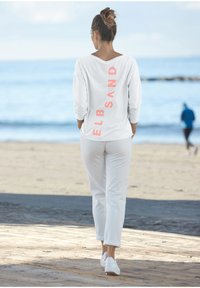 Weißes Langarmhemd mit pinkem vertikalem Text "ELBSAND" auf dem Rücken, kombiniert mit weißen Hosen und weißen Sneakers, vor einem Strandhintergrund.