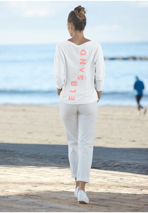 Weißes Langarmhemd mit pinkem vertikalem Text "ELBSAND" auf dem Rücken, kombiniert mit weißen Hosen und weißen Sneakers, vor einem Strandhintergrund.